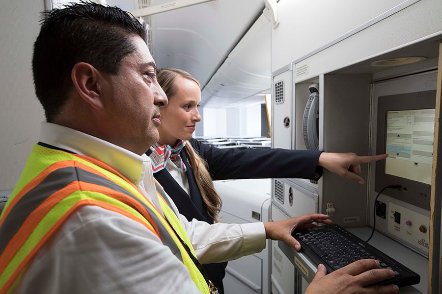 Two people, one in a high-visibility vest and the other in a navy uniform, interact with a monitor on an aircraft